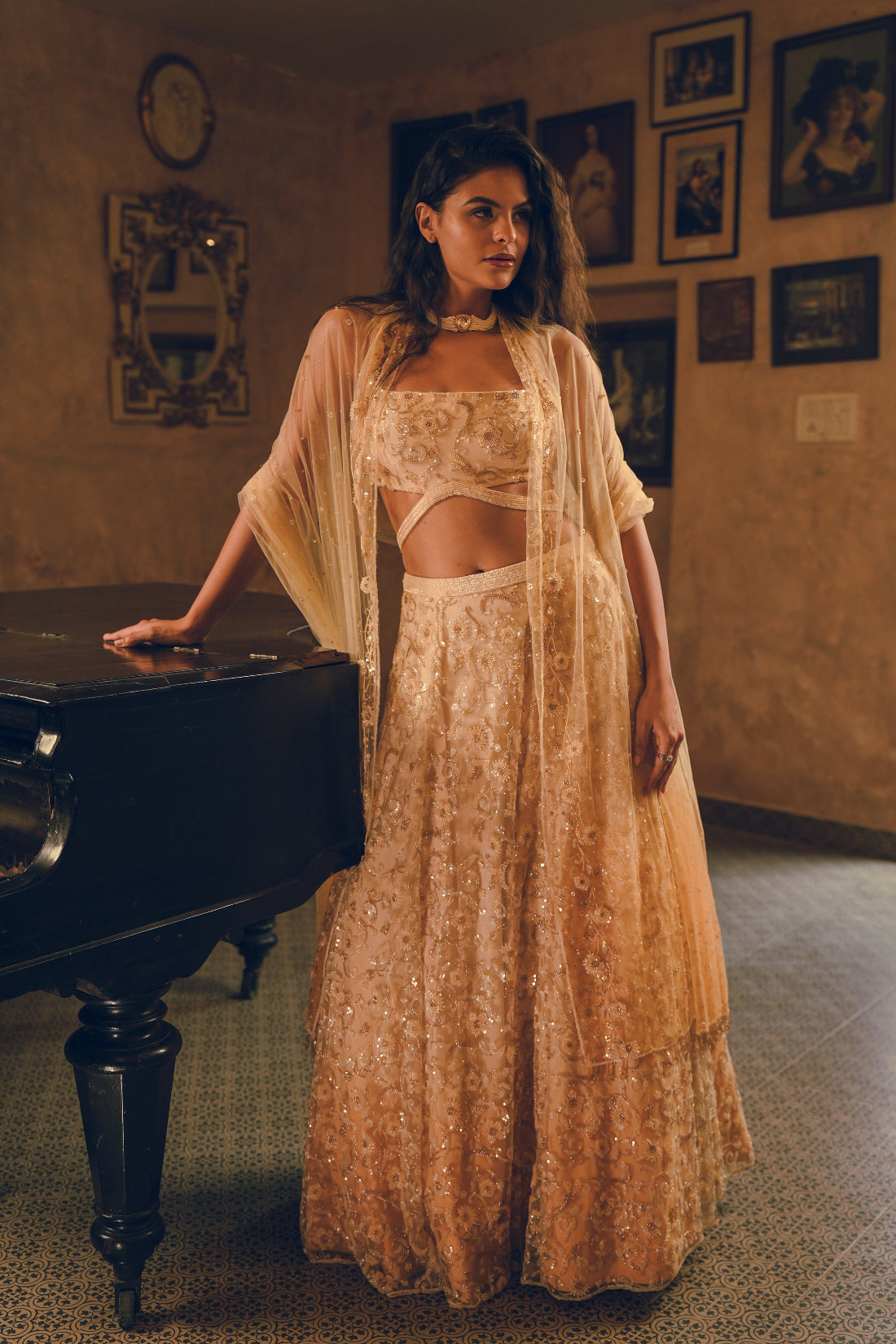 Woman in a long, flowing dress standing next to a piano in a room with framed pictures on the wall.